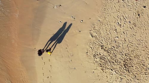 Two girls casually walking down the shore of a tropical beach, with shallow waves crashing next to t