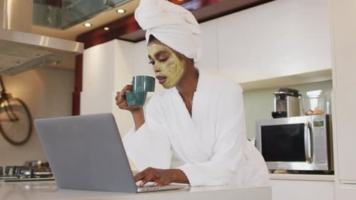 African american attractive woman with applied face mask drinking coffee and using laptop in kitchen