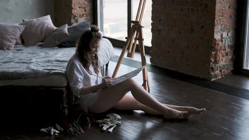 Painter Paints a Picture Sitting on the Floor in a Home Studio. Girl in a Man's Shirt