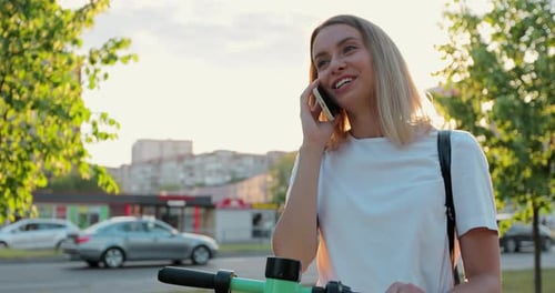 Gros plan d'une femme heureuse debout avec un scooter électrique moderne parlant au téléphone en ville