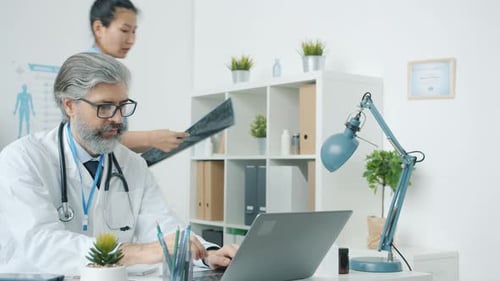 Doctor Using Laptop Then Discussing MRI Scans with Nurse Working in Modern Hospital Office