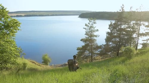 Hiker with Backpack Stands on Grassy Hillside Overlooking Lake