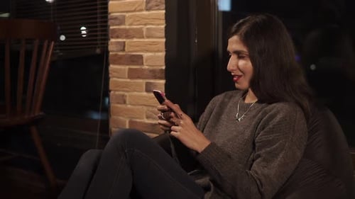 Brunette sitzt abends in einem Café auf einem Sitzsackstuhl und benutzt Smartphone