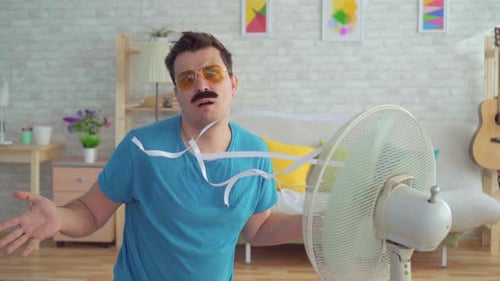 Divertido joven frente a un ventilador eléctrico que funciona escapa del calor en el apartamento