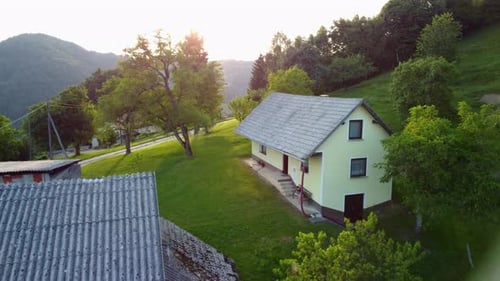 Flying toward small countryside vacation house in hills between green nature with sun in background.