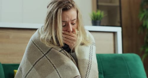 Kranke junge Frau zu Hause auf dem Sofa mit einer Erkältung, die sie mit einer Decke bedeckt und sich die Nase putzt