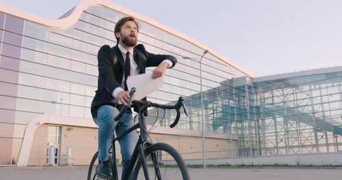 Empresario que monta en bicicleta cerca de una moderna construcción urbana de vidrio y arroja papeles comerciales