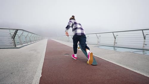 Young Guy in a Checkered Black and White Shirt Rides on a Board Across the Bridge