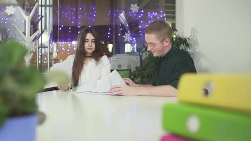 Pretty Girl and Young Man in Glasses Sitting at the Table in Modern Office Reading Papers Discussing