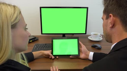 Two office workers sit at a desk and look at a computer and a tablet with green screens
