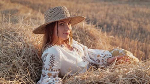 Pretty Woman in Straw Hat and Embroidered Blouse Smiling Lying on Hay in Countryside at Sunset