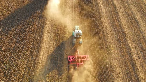 Top View Tractor with Harrow System Plowing Ground on Cultivated Farm Field Pillar of Dust Trails