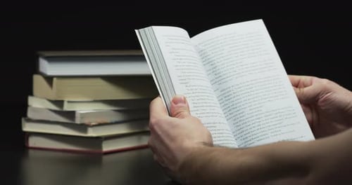 Person Reads Paperback Book With Stack of Books