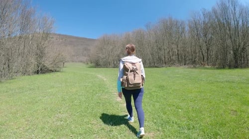 Young Woman Traveler with a Backpack Walks Along the Trail Past Meadows and Forests