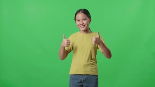 Fröhliches junges asiatisches Mädchen zeigt eine Daumen-Hoch-Geste, während es im Greenscreen-Studio steht