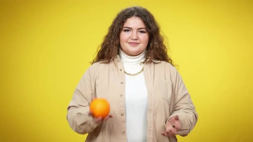 Charming Plussize Caucasian Brunette Woman Standing at Yellow Background Juggling and Smelling