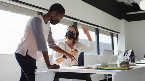 Two diverse female colleagues wearing face masks looking at laptop and discussing in office
