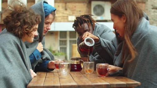 Friends in Blankets Sit Outside the Cafe and Pour Tea in Cups
