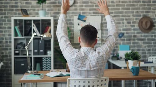 Back View of Young Man Stretching Arms Relaxing Sitting in Office at Desk Alone