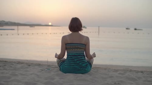 Femme brune des années 30 en robe assise en position du lotus et méditant sur la plage au bord de la mer