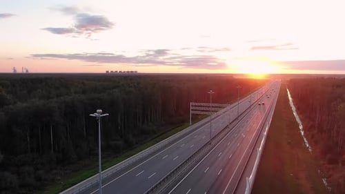 Wide Road Among Forests and Driving Cars at Orange Sunset
