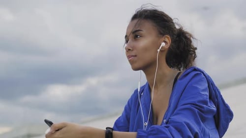 Young Woman Pauses with Phone and Earbuds Outdoors
