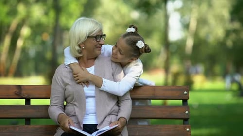 Little Girl Running to Grandmother Reading Book in Park, Hugging and Kissing Her
