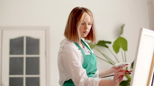 Young Woman in an Apron Paints on Canvas with Brush While Resting After Work Front View