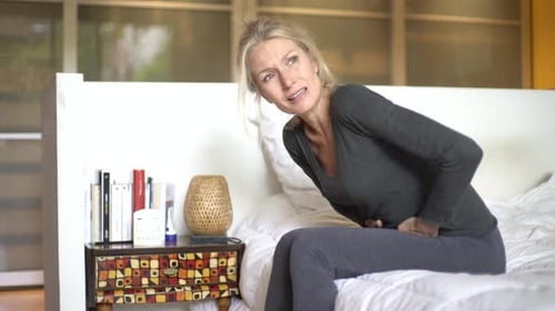 Woman Sits with Abdominal Pain in Bedroom