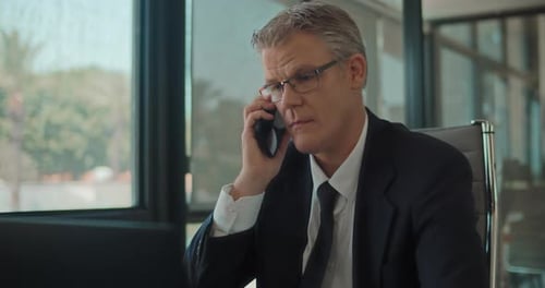 Close up of a businessman in formal wear and glasses answering a phone while working on a laptop