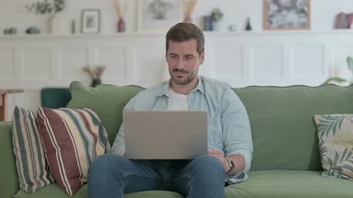 Young Man with Laptop Reacting to Loss on Sofa