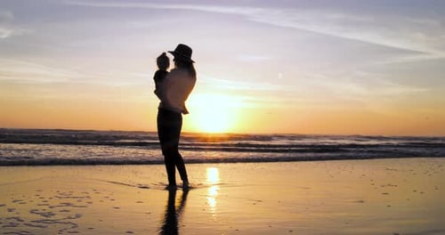 A woman enjoys a stunning and colorful sunset with her child at the beach in 4k.