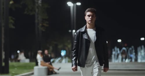 Young Man Walking in Urban Park Late in Evening, People and Fountain on Background, Slow Motion