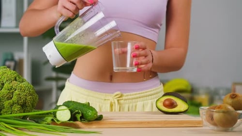 Young Woman Pouring Green Detox Smoothie Into Glass From Blender and Putting on Table in the Kitchen