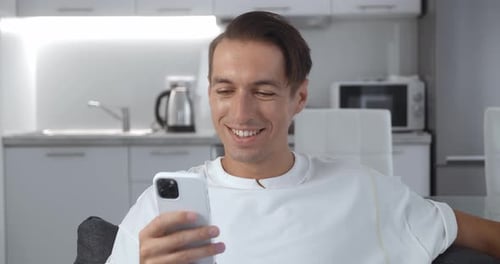 Close Up of Cheerful Young Man Sitting on the Sofa Using Smartphone Smile in the Modern Apartment