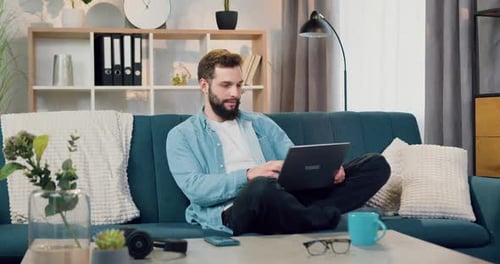 Homme barbu assis dans une pose de lotus sur le canapé à la maison et travaillant sur un ordinateur