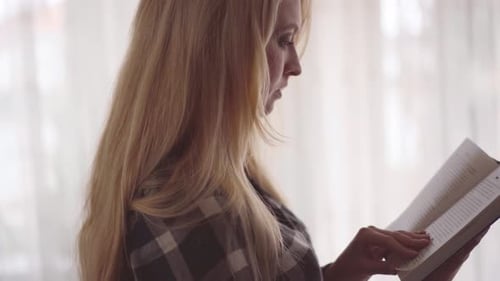 Side View of Young Woman Reading Interesting Book in the Room in Front of Large Window Close Up