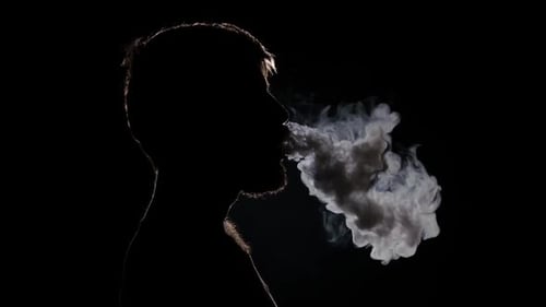 exhaling cigarette smoke woman smokes thin cigarette, exhaling smoke. Female hand with smoking cigarette on blurred green park background Stock Photo Exhaling cigarette smoke hi-res stock