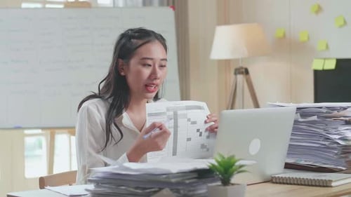 Asian Woman With Documents Explaining Work On Paper While Having Video Call On Laptop At The Office