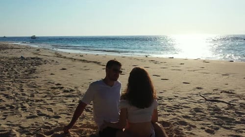 Cute couple sitting on sand of quiet exotic beach on a bright sunset reflected over seashore full of