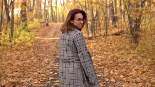 A Young Woman in Glasses and a Coat Walks in the Autumn Forest. Yellow Leaves Around. Slow Motion