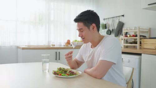 Asian attractive handsome male eating green salad in kitchen at home.