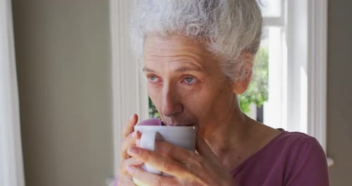 Close up of caucasian senior woman drinking coffee while looking out of the window at home