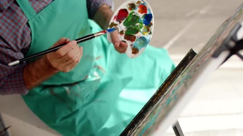 Man Artist Works in Workshop Zeichnen auf Leinwand mit Pinsel und hält Farbpalette in der Hand Top