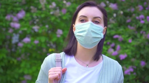 Portrait of a Young Woman in a Medical Mask on the Background of Flowering Plants in the Park
