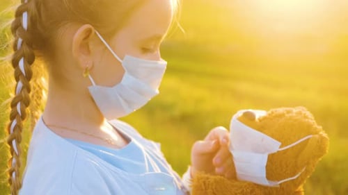 Weinendes Mädchen in medizinischer Schutzmaske mit einem Teddybär bei Sonnenuntergang