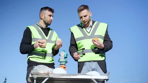 Medium Shot Two Caucasian Workers Discussing Blueprint Eating Lunch on Construction Site Outdoors