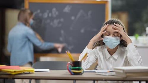 Portrait of Exhausted African American Girl in Covid19 Face Mask Sitting at Desk in Classroom
