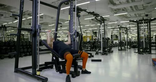 Girl Lifts Up Barbell Lying on Bench By New Trainer in Gym