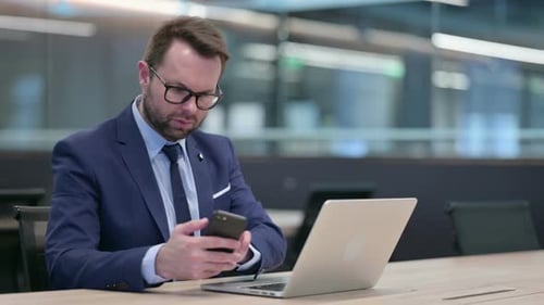 Man Works on Laptop and Phone in Office